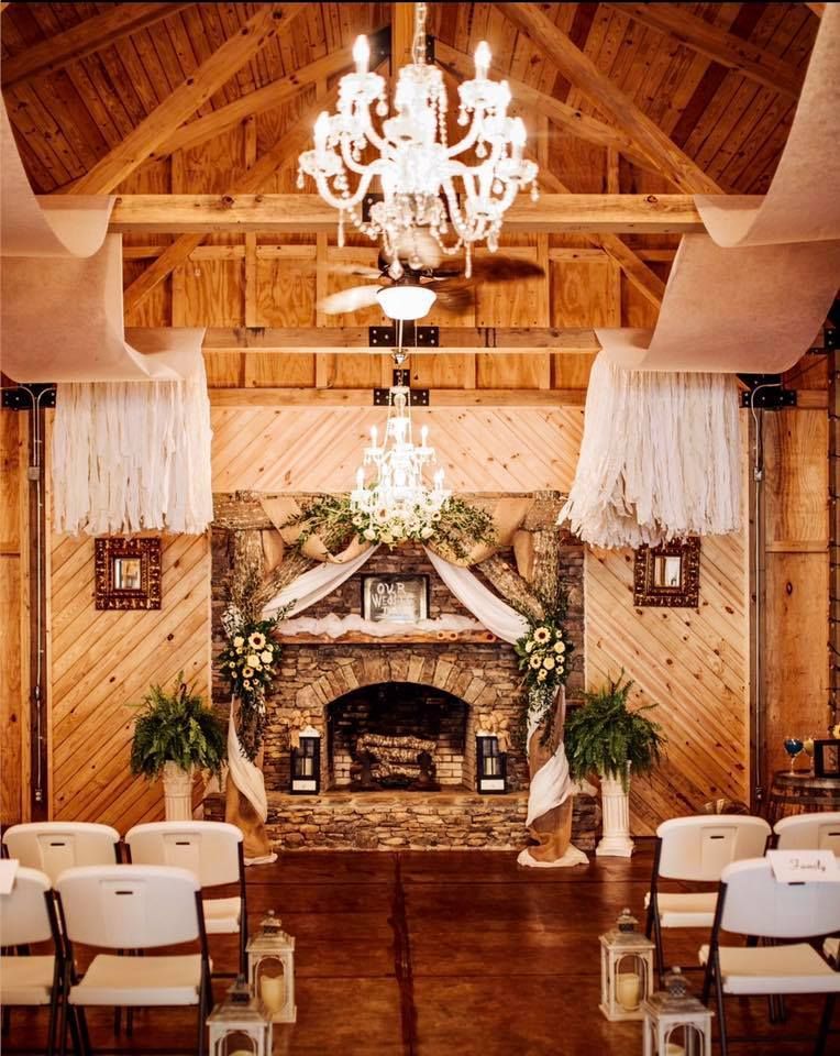 Elegant barn-style wedding venue with chandeliers, floral arch, fireplace, and rows of white chairs