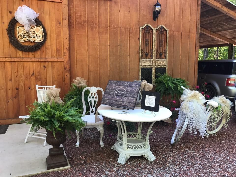 Outdoor rustic porch display with white table, chairs, potted ferns, and floral arrangements against a wooden wall