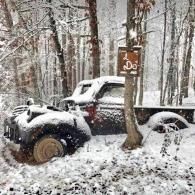 Snow-covered off-road vehicle parked in a snowy forest beside a tree with a sign.