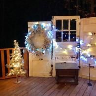 Holiday-decorated porch with lit Christmas tree, wreath, string lights, and white shutters at night