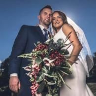 Newlyweds posing outdoors, smiling with a large bouquet and veil against a blue sky