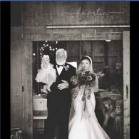 Bride and groom stand at a doorway, holding bouquets and a doll, in a black-and-white photo.