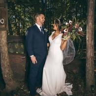Wedding couple posing outdoors in a wooded setting, bride in white dress holding bouquet, groom in navy suit