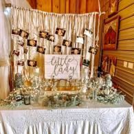 Rustic dessert table with “little lady” sign, hanging photos, and glass jars in a warm wood-paneled room