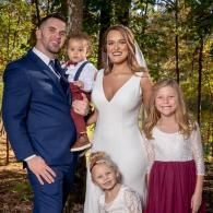 Family posing outdoors in formal attire, with a man, woman, toddler, and two young children in autumn trees