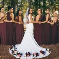 Bride in a white gown with bridesmaids in burgundy dresses, standing in a wooded outdoor setting.