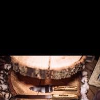 Two knives in a wooden case beneath a round tree stump, with bark and feathers around them.