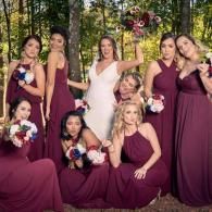 Seven people in burgundy dresses pose outdoors in a wooded setting, holding bouquets.