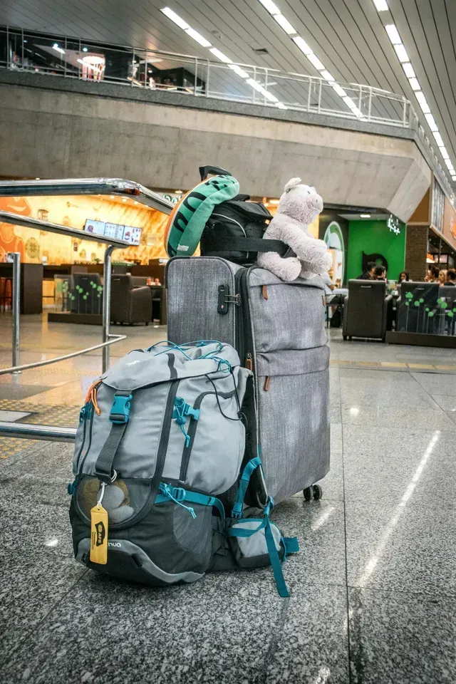 Pilha de bagagens em um aeroporto: mochila, malas e bichos de pelúcia em um chão, perto de grades de metal e uma loja verde.
