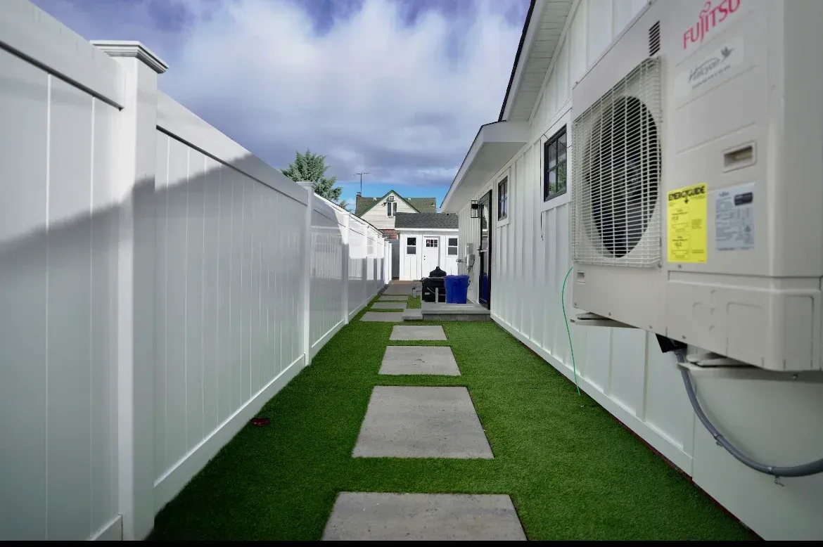 Narrow outdoor walkway with stepping stones, flanked by white fence and building with HVAC unit. Green grass and blue sky.