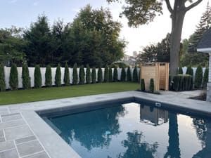 A rectangular pool reflects the sky, surrounded by a stone patio and bordered by a row of evergreens.