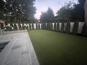 A backyard with a rectangular lawn, stone patio, and tall green trees lining a white fence.