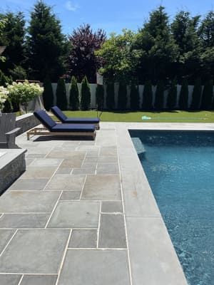 Poolside patio with blue lounge chairs on gray stone pavers; pool on right, green lawn in background.