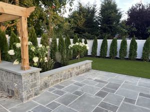 Patio with stone pavers, low wall, arbor, and green lawn with evergreen trees against a white fence.
