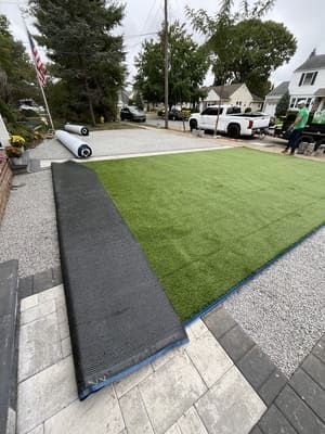 Rolls of artificial turf being installed on a patio, gray pavers and gravel surround the green grass.