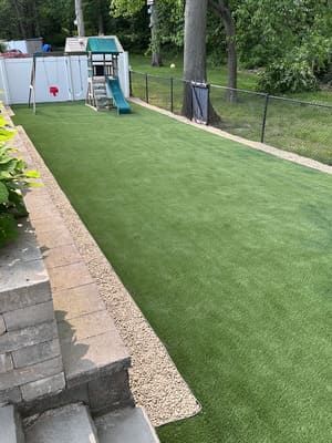 Green artificial turf play area with swing set, slide, bordered by stone and fence.