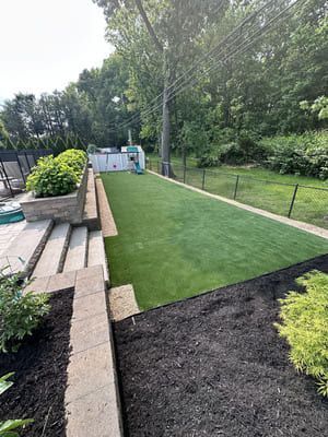 Backyard with artificial turf, bordered by black mulch, stone steps, and a black fence.