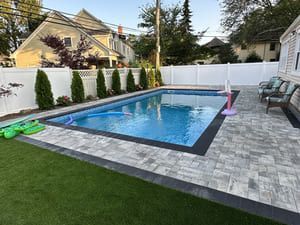 Backyard with a rectangular swimming pool, patio, white fence, and green lawn.