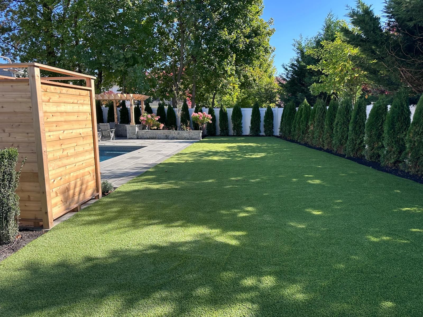Backyard with green artificial turf, wooden fence, trees, and white fence.