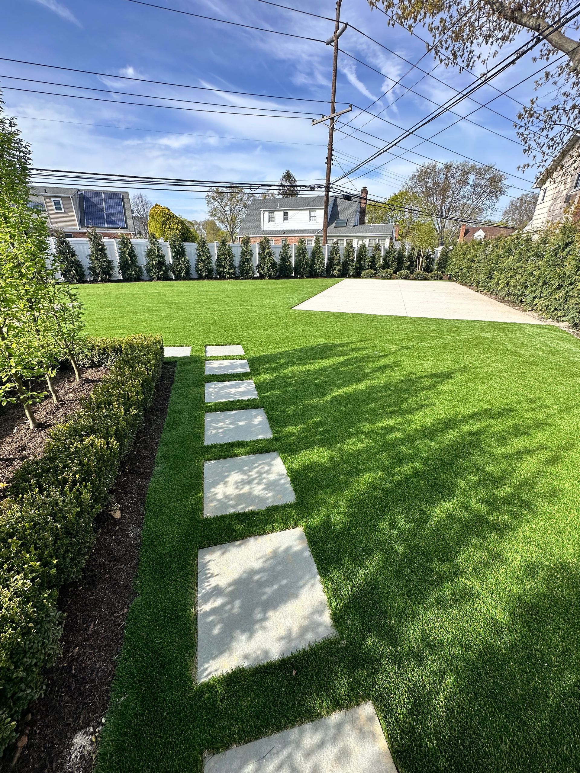Square stone path through a green lawn, edged by hedges. Clear blue sky overhead.