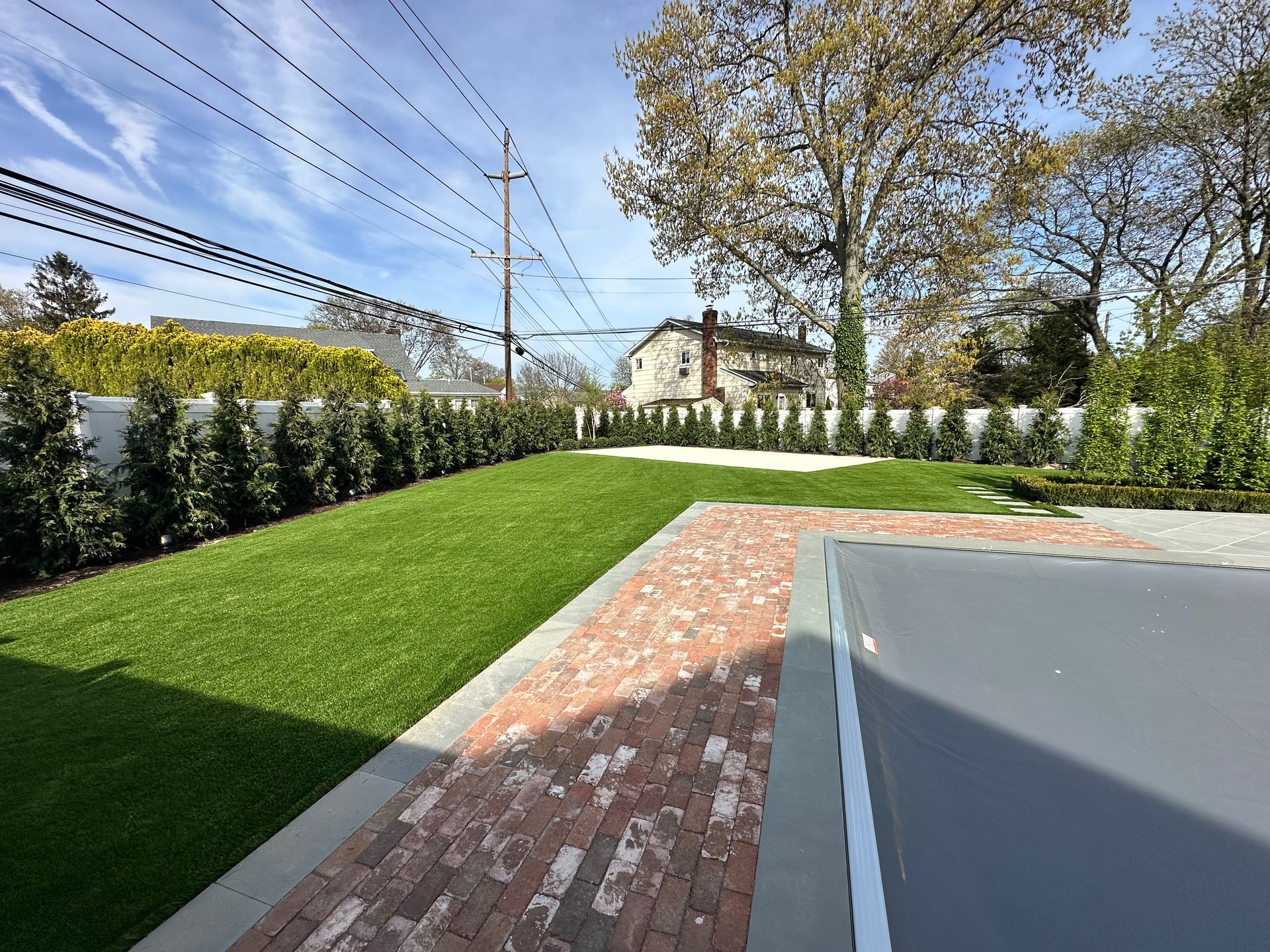 Backyard with brick path, green lawn, white fence, and trees under a blue sky.