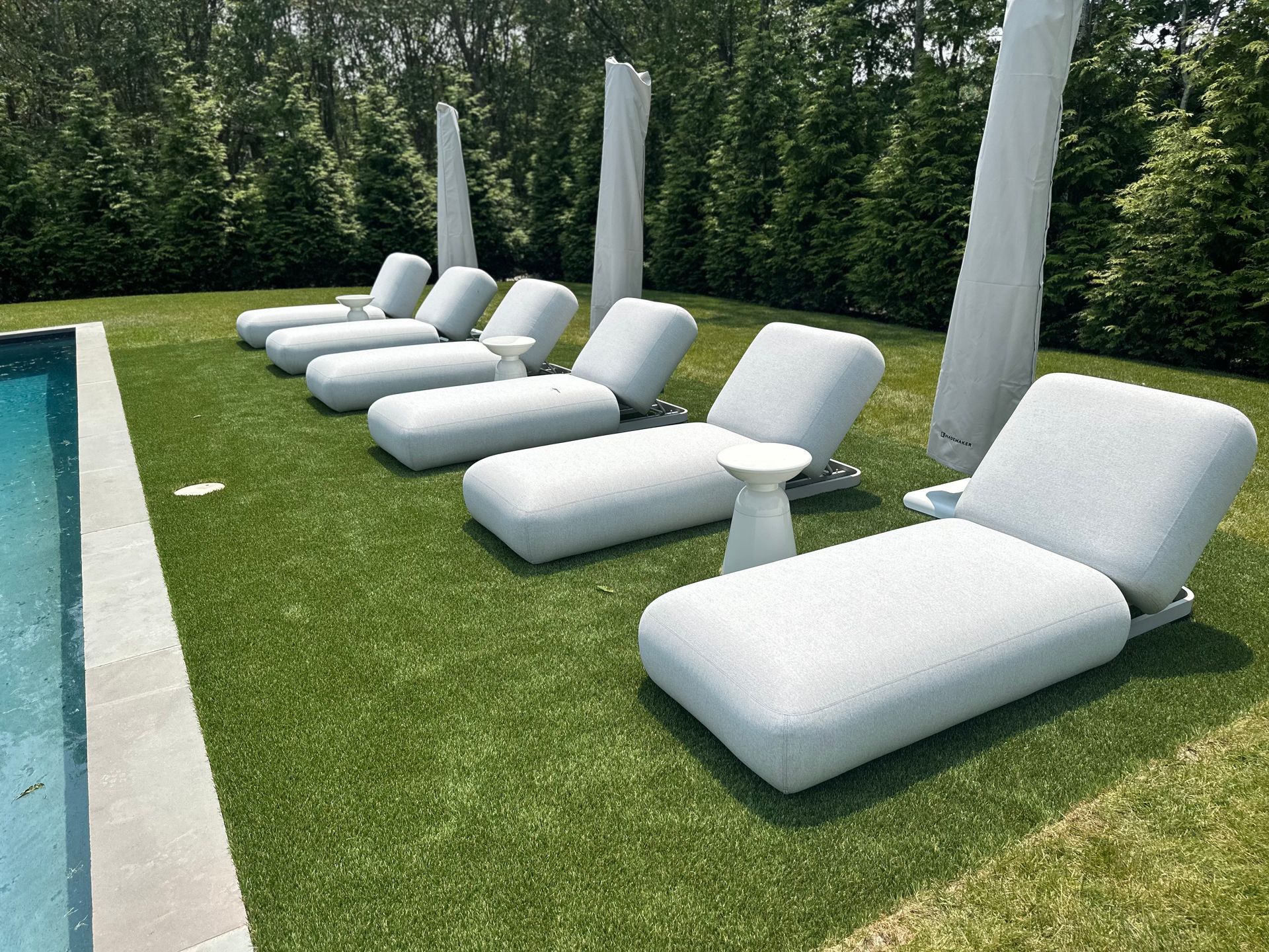 Row of white chaise lounges with small side tables near a pool, on green turf, with tall shade structures and a backdrop of green trees.