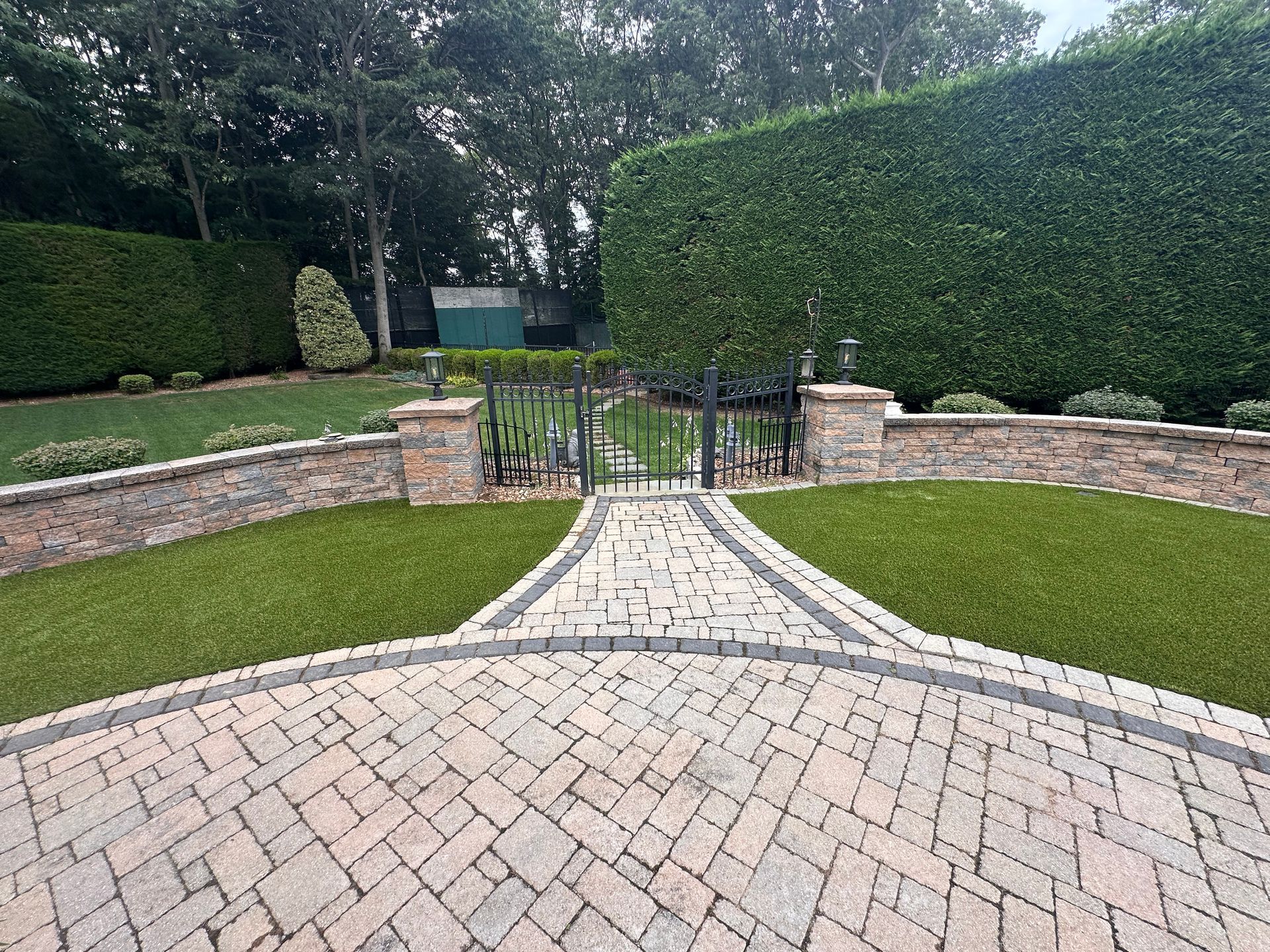Brick driveway leading to a gate, surrounded by green lawns, hedges, and a stone wall.