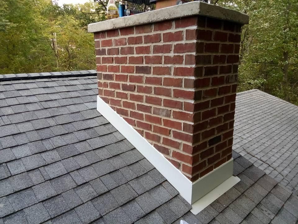 Brick chimney on a gray shingled roof with white flashing, against a wooded background