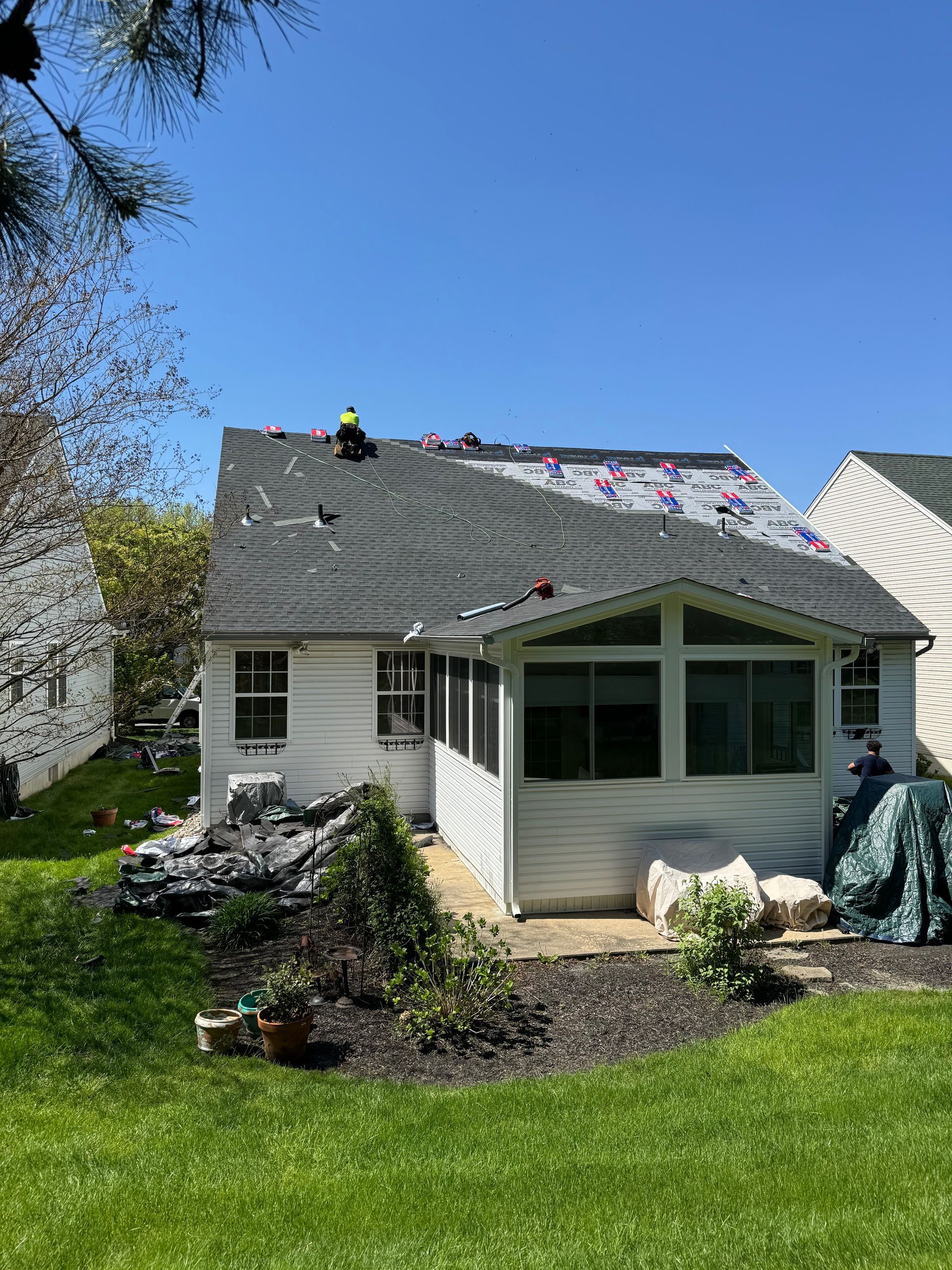 House with roofers installing dark shingles on a sunny day, with lawn and garden in front.