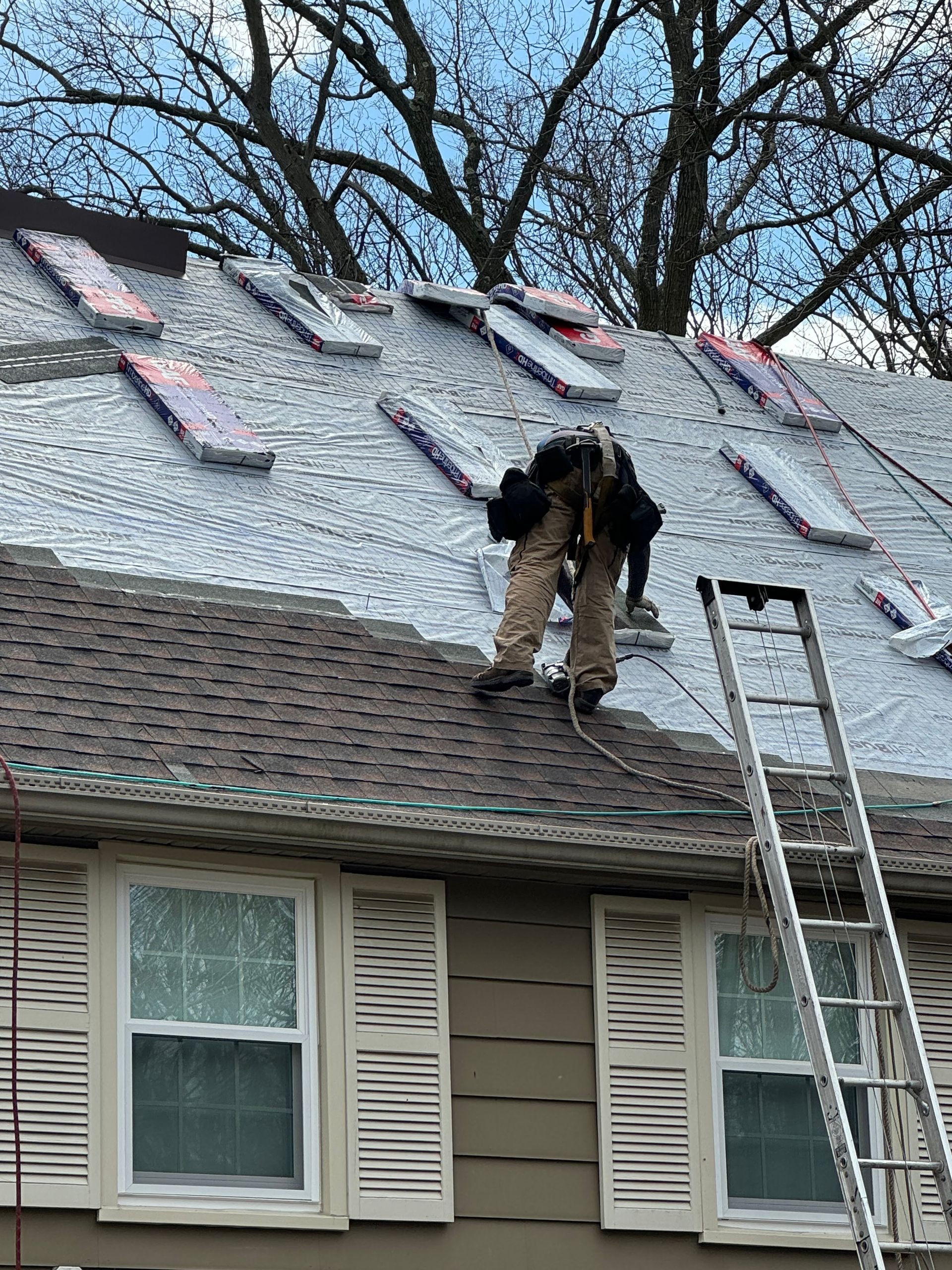 Worker on a house roof replacing shingles beside a ladder.