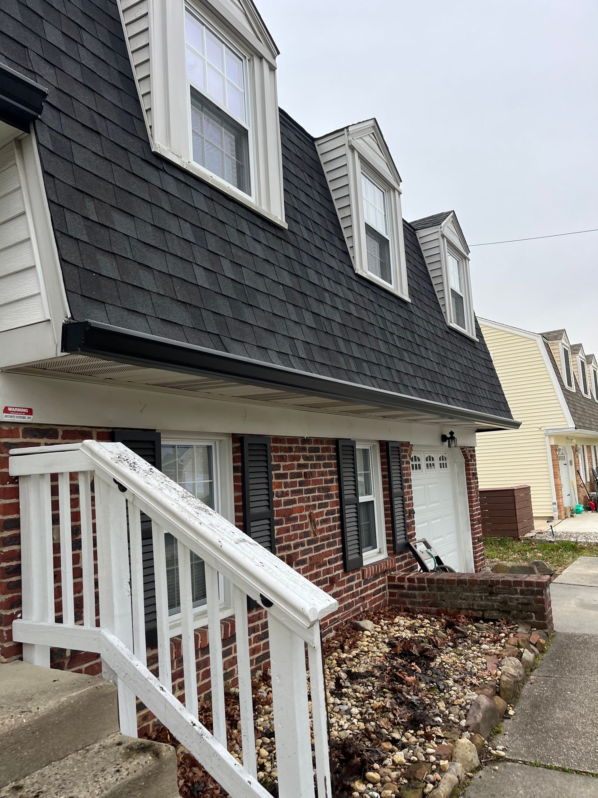Brick townhouse with dark shingle siding, white stairs, and dormer windows beside a sidewalk.