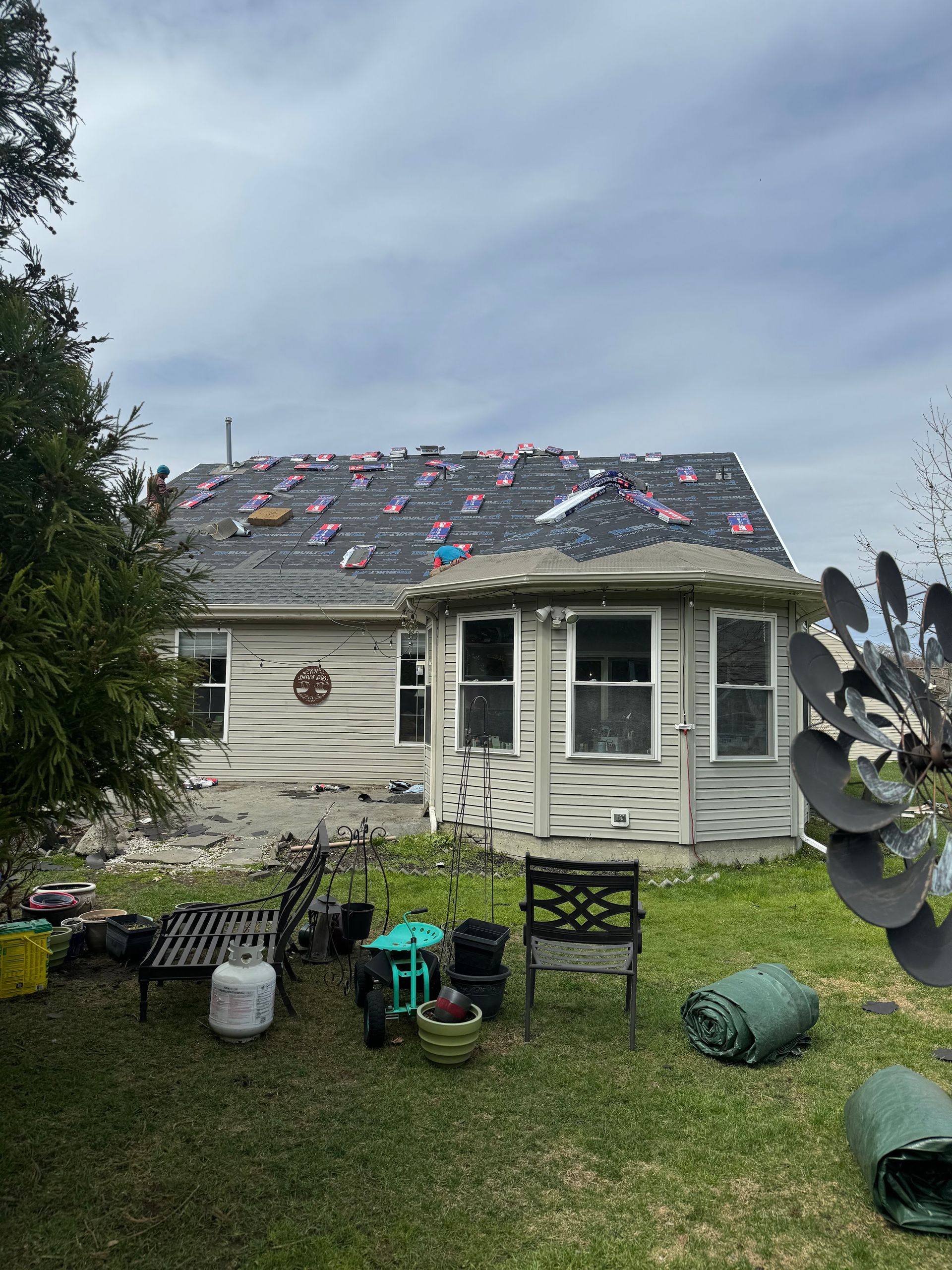 Small white house with damaged roof covered in colorful patches, grassy yard, and scattered outdoor chairs.