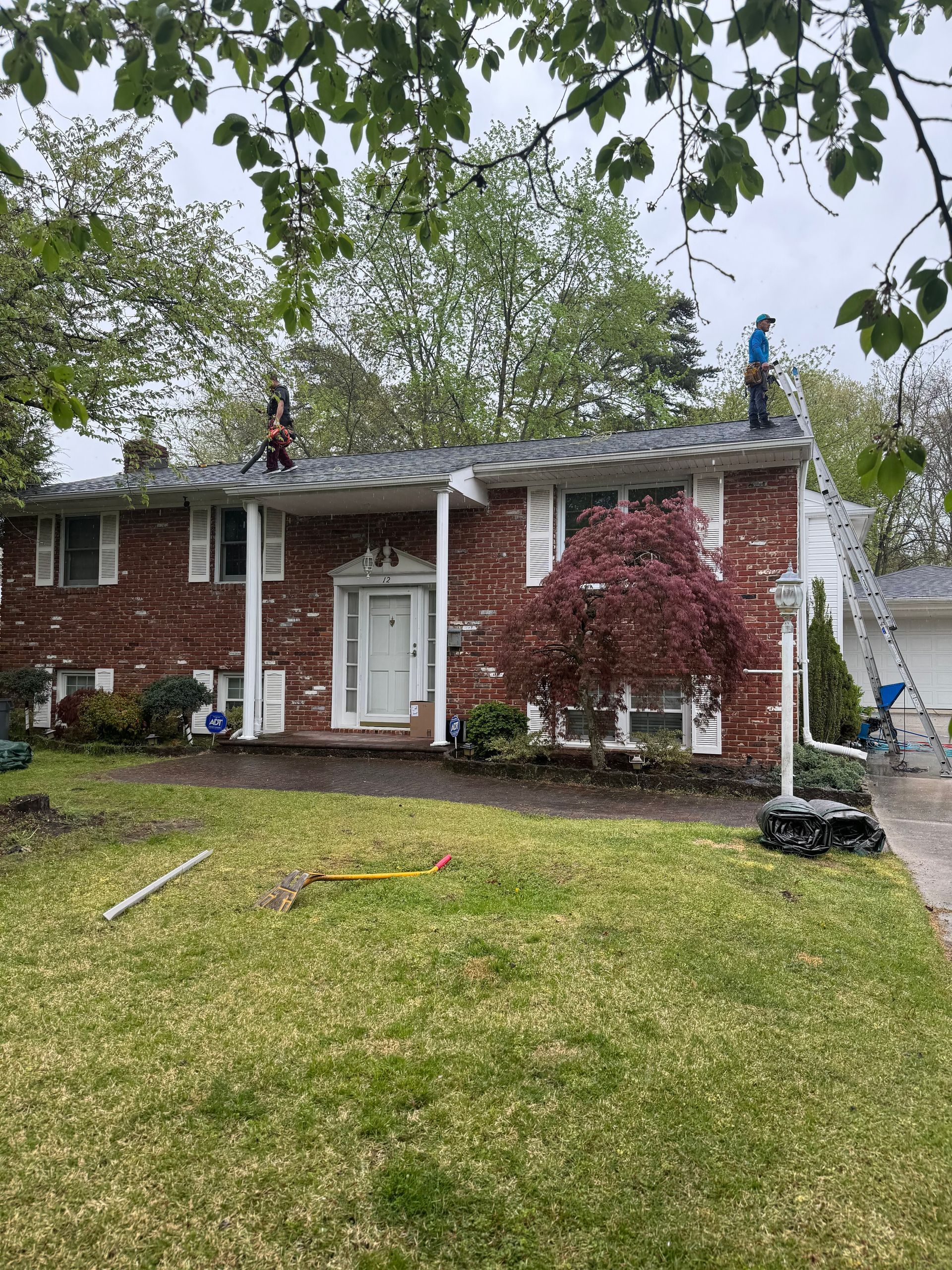 Brick house with white columns, two workers on the roof, and a ladder on the right in a front yard.