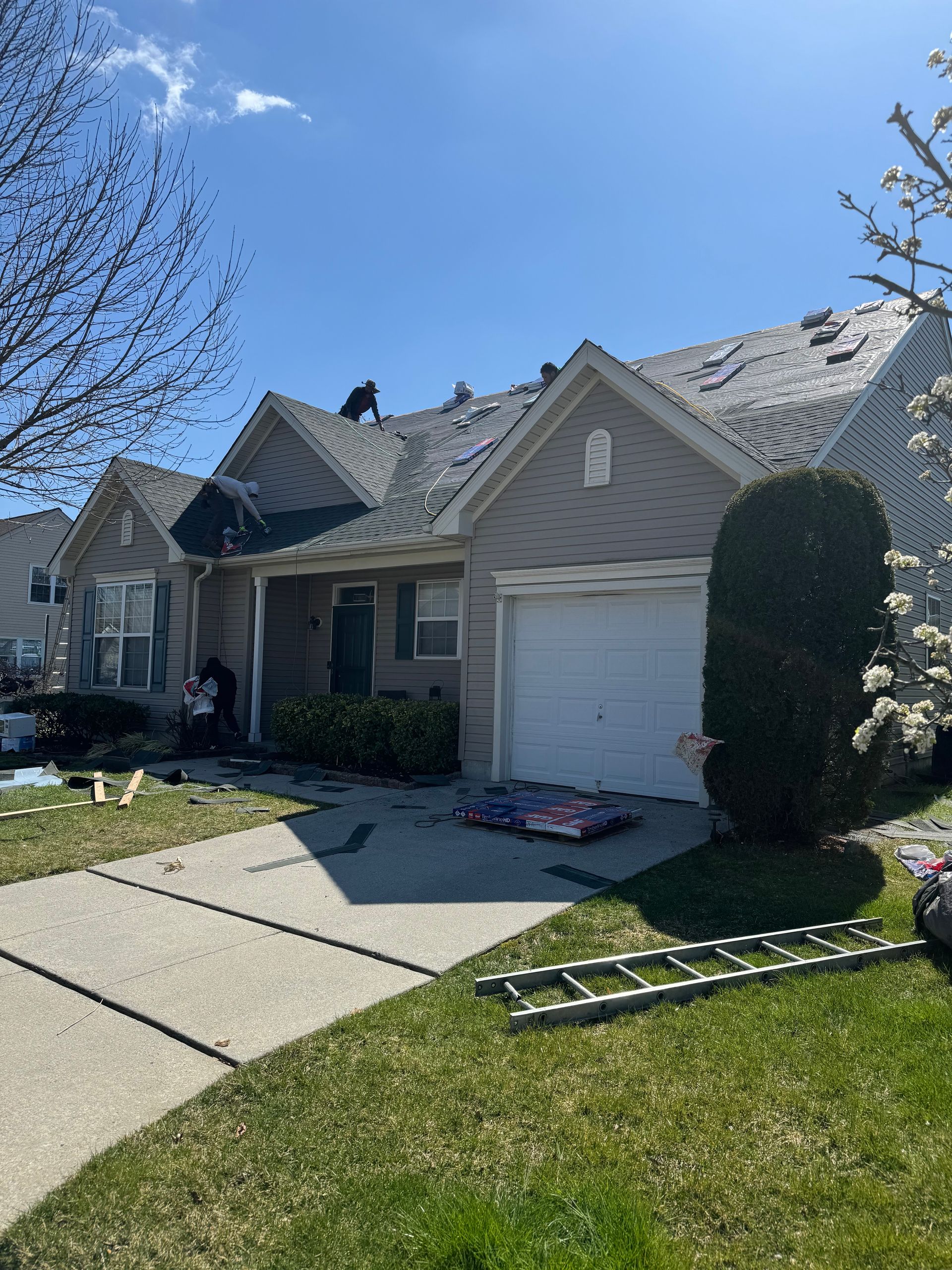 Suburban house with a damaged roof, ladder on the lawn, and a garage door in the driveway.