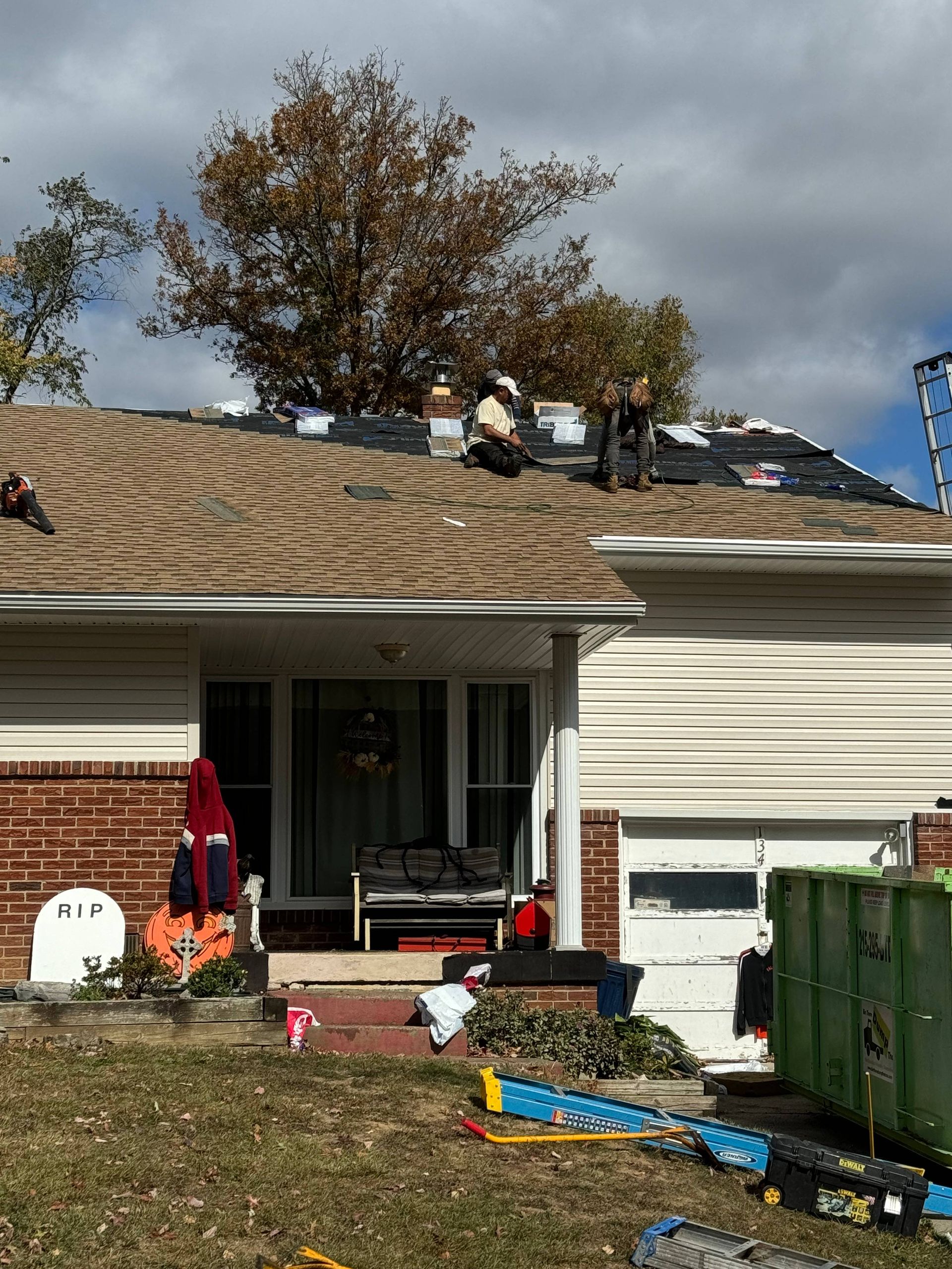 Home roof with storm damage and workers on top, yard cluttered with bins and supplies in front