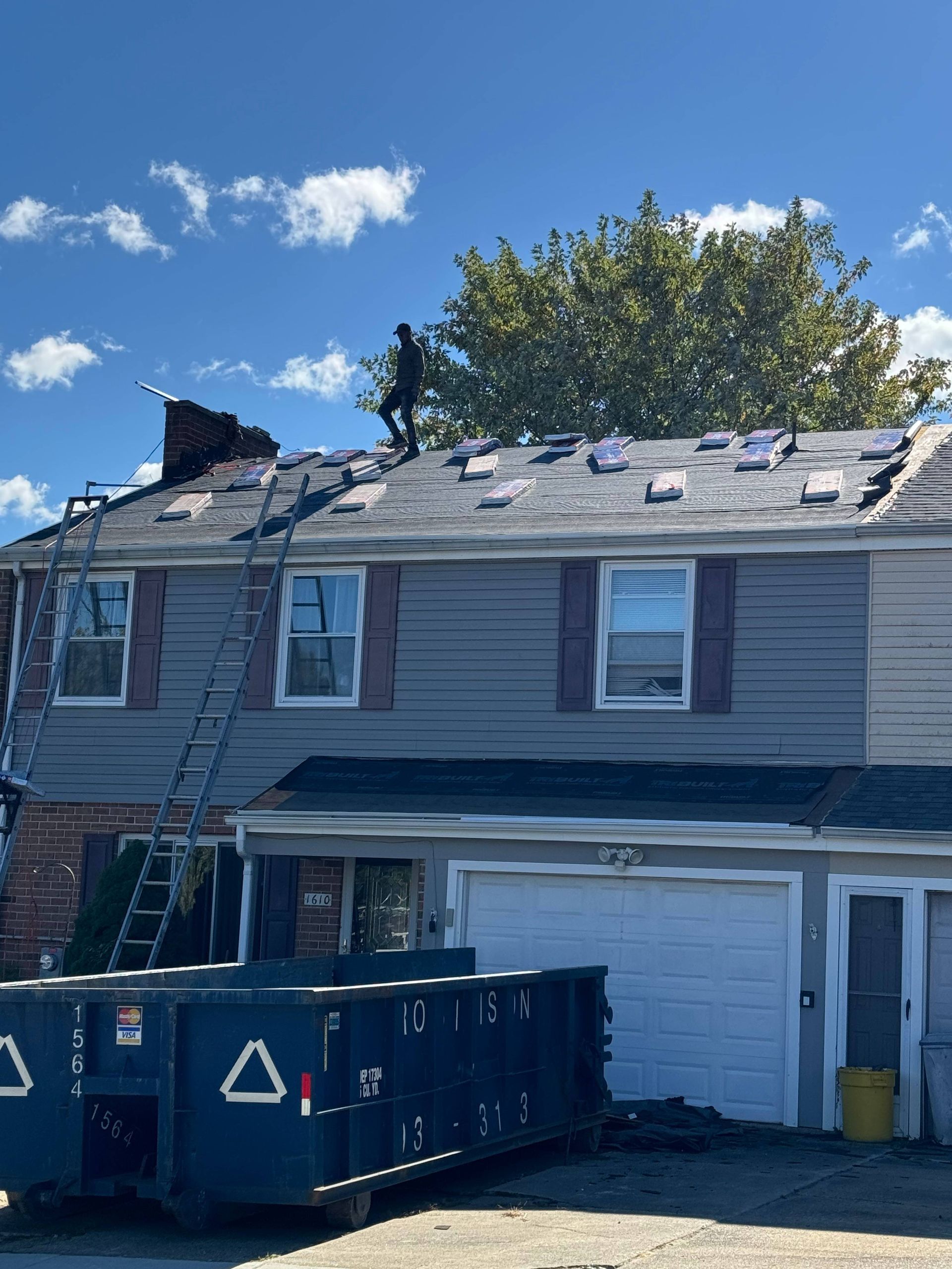 Damaged house roof with exposed sheathing, dumpster in front, and ladder leaning against the side.