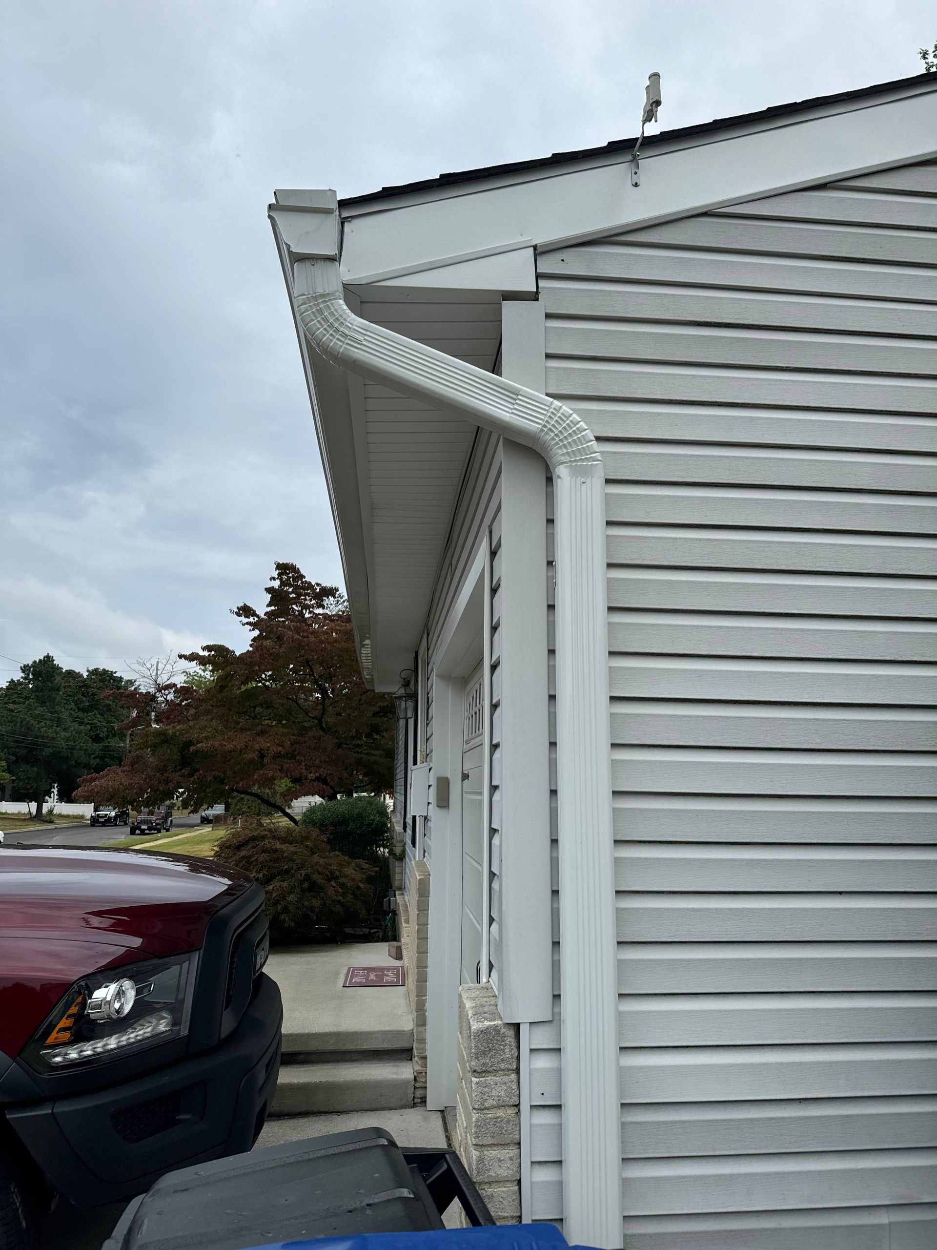 House siding with white gutter and downspout beside a parked maroon SUV under cloudy sky