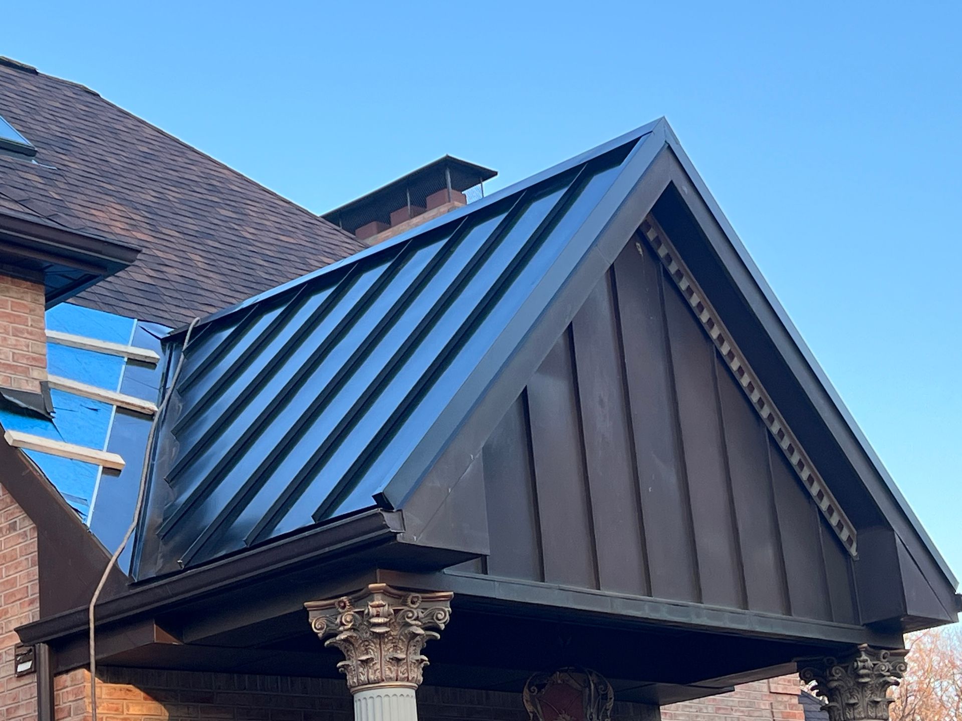Close-up of a gabled roof with dark metal siding and ornate stone column detail under blue sky