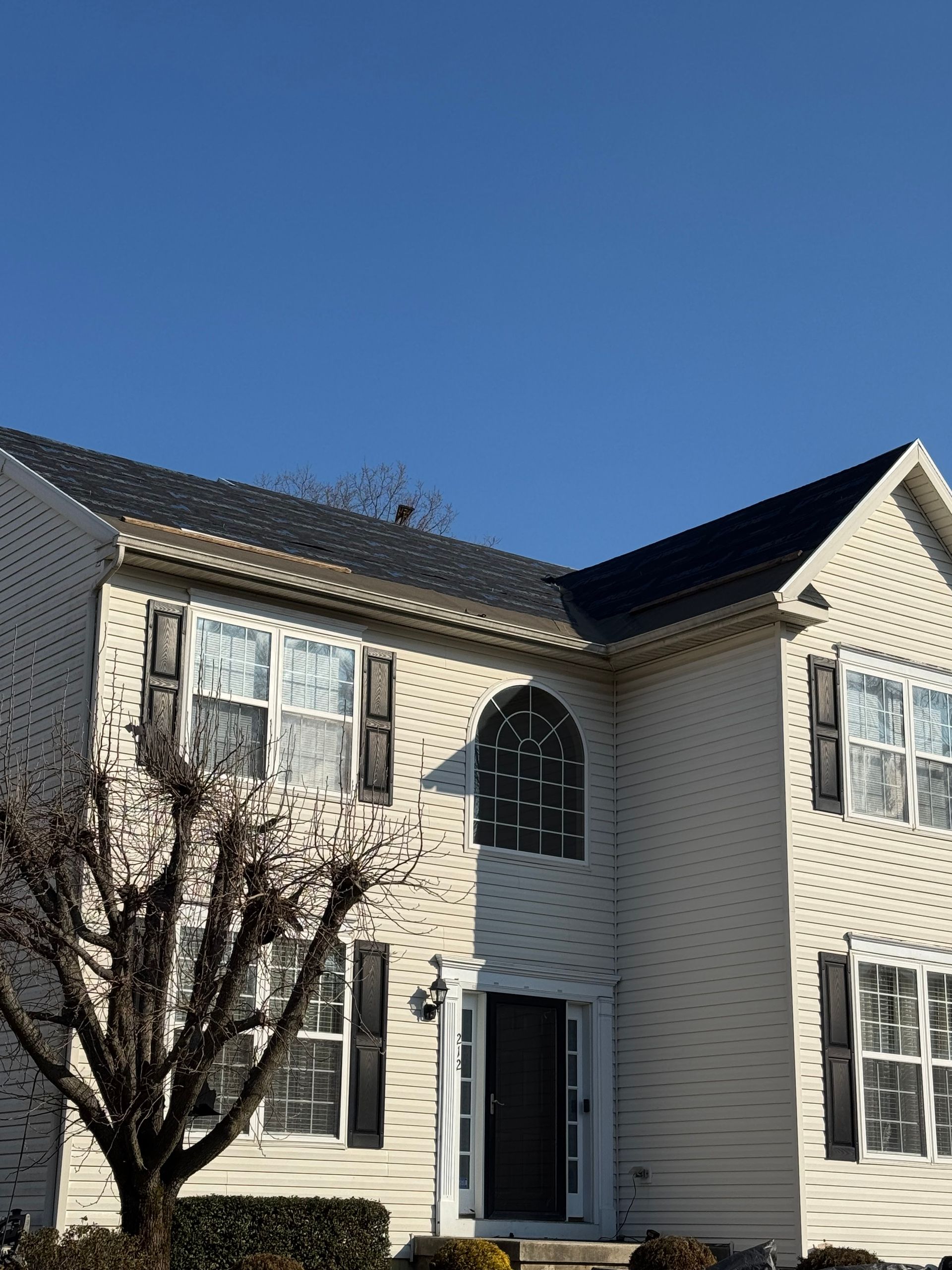 Two-story beige house with white shutters, dark roof, and a front door under a clear blue sky