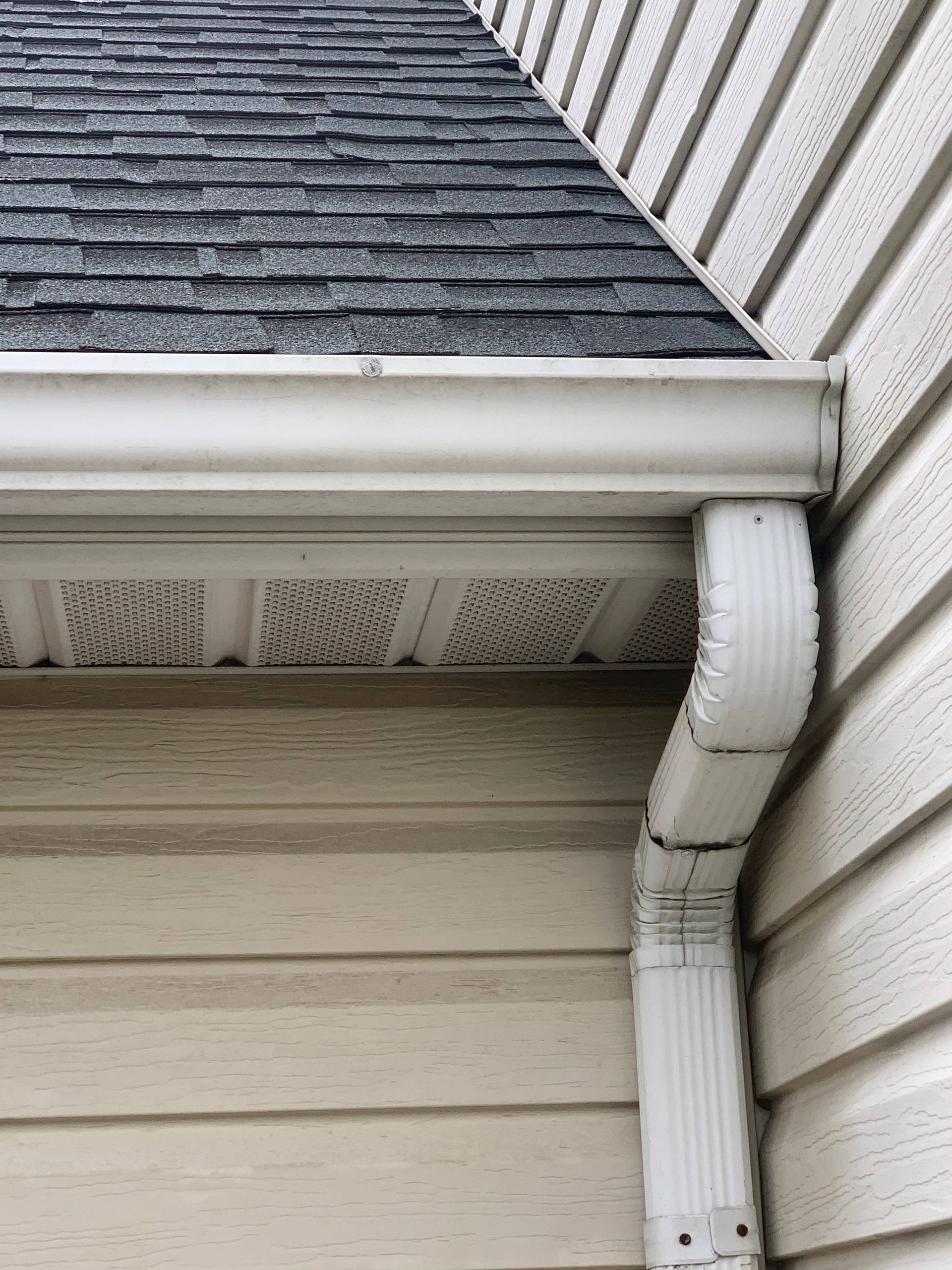 House gutter and downspout corner beneath shingled roof and vinyl siding