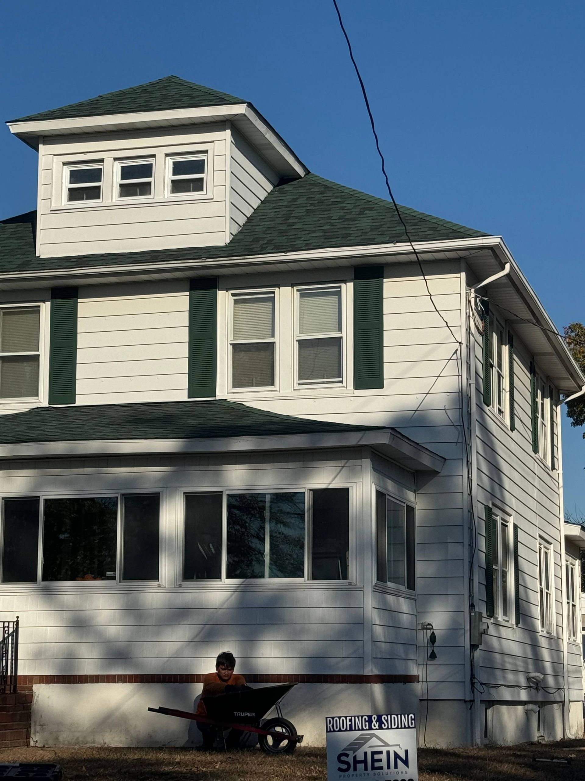 White two-story house with dark green roof and shutters, front porch, and a person seated on the lawn.