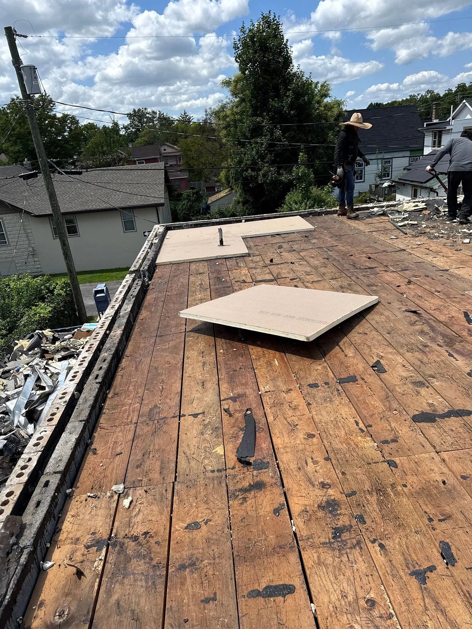 Rooftop with exposed plywood and roof materials, showing ongoing repair work near trees and houses.