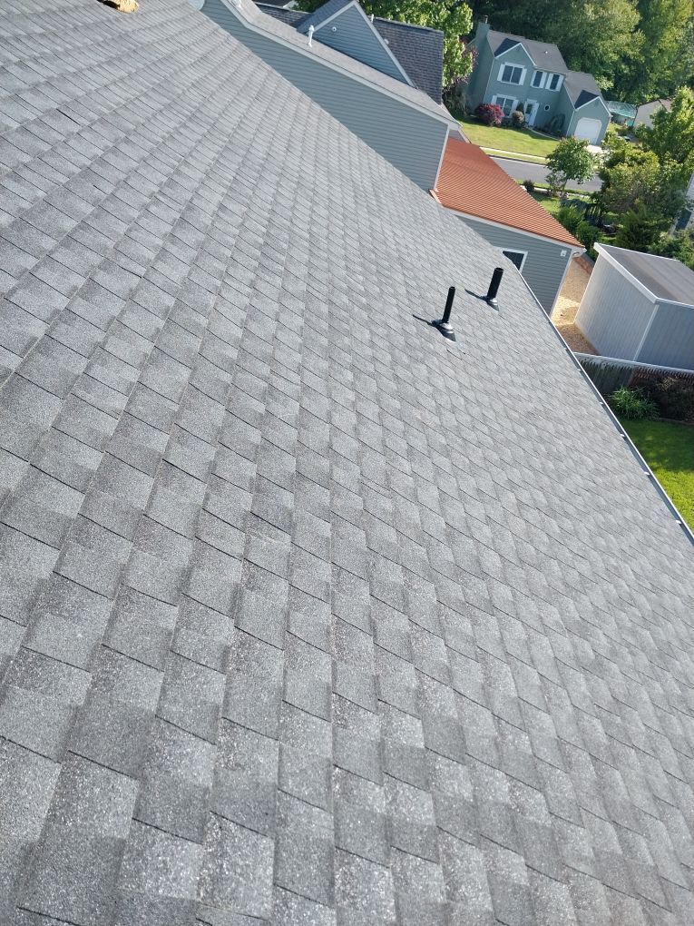 Tilted view of a gray shingled roof with two black vent pipes and nearby houses below