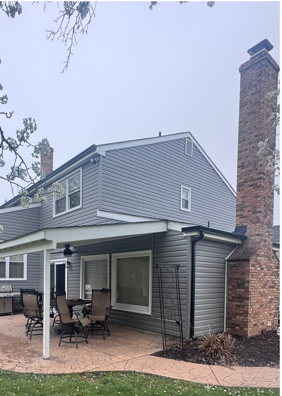 Gray-sided house with a covered patio, outdoor chairs, and a tall brick chimney