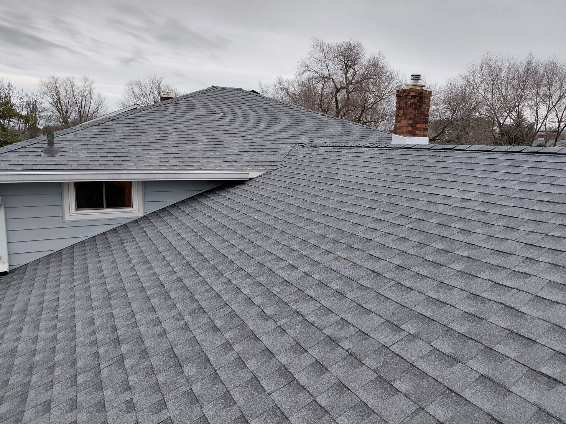Snow-covered roof with a brick chimney and bare trees in the background