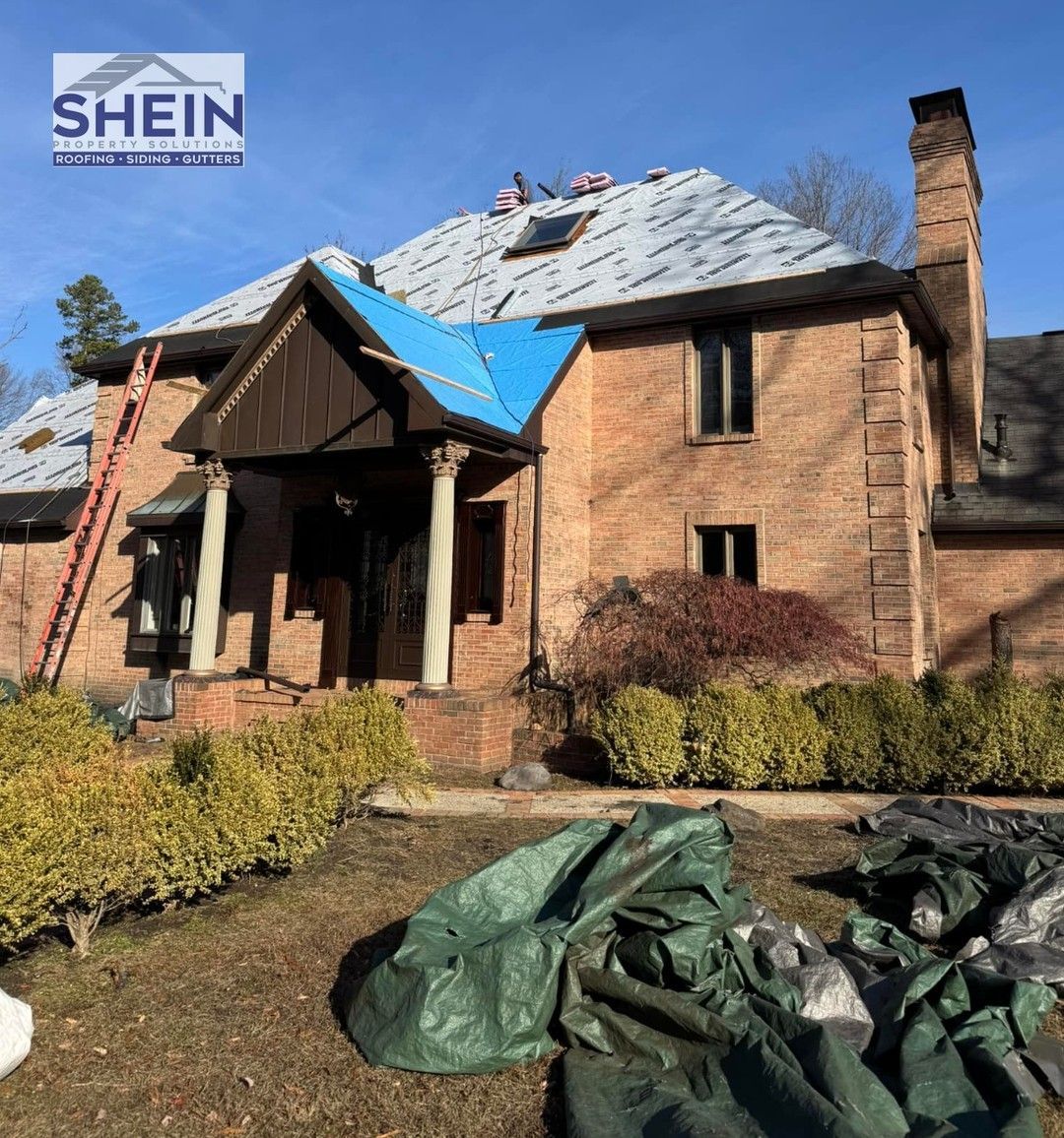 Damaged brick house with torn roof tarp and debris in front yard under a clear blue sky