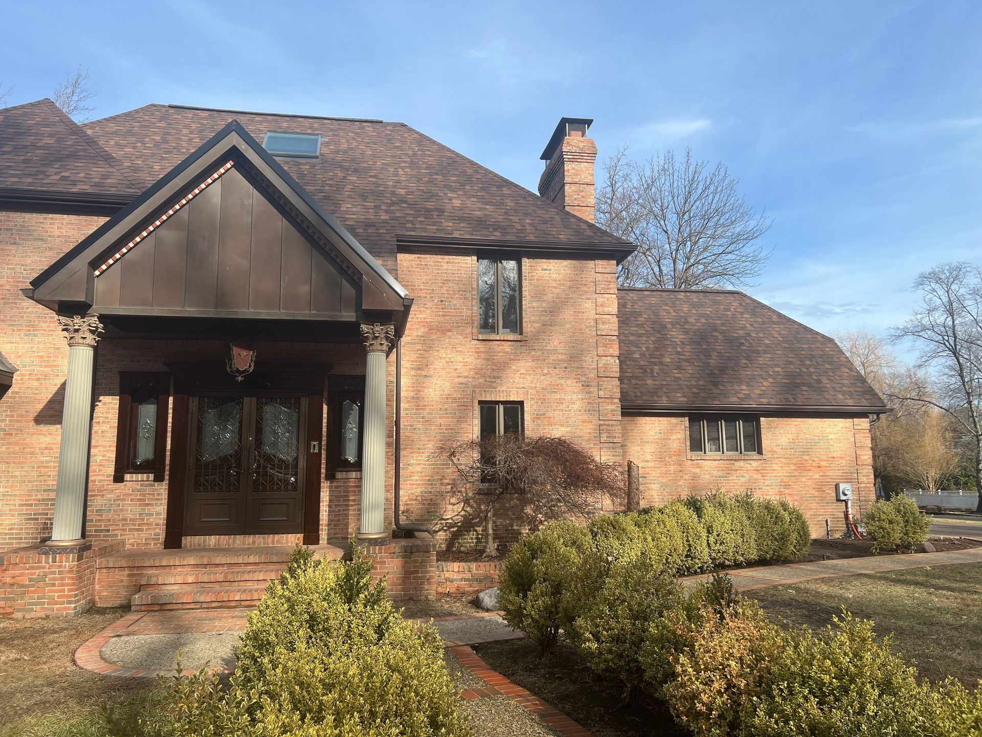 Brick house with a covered front porch, shingled roof, and trimmed bushes on a sunny day