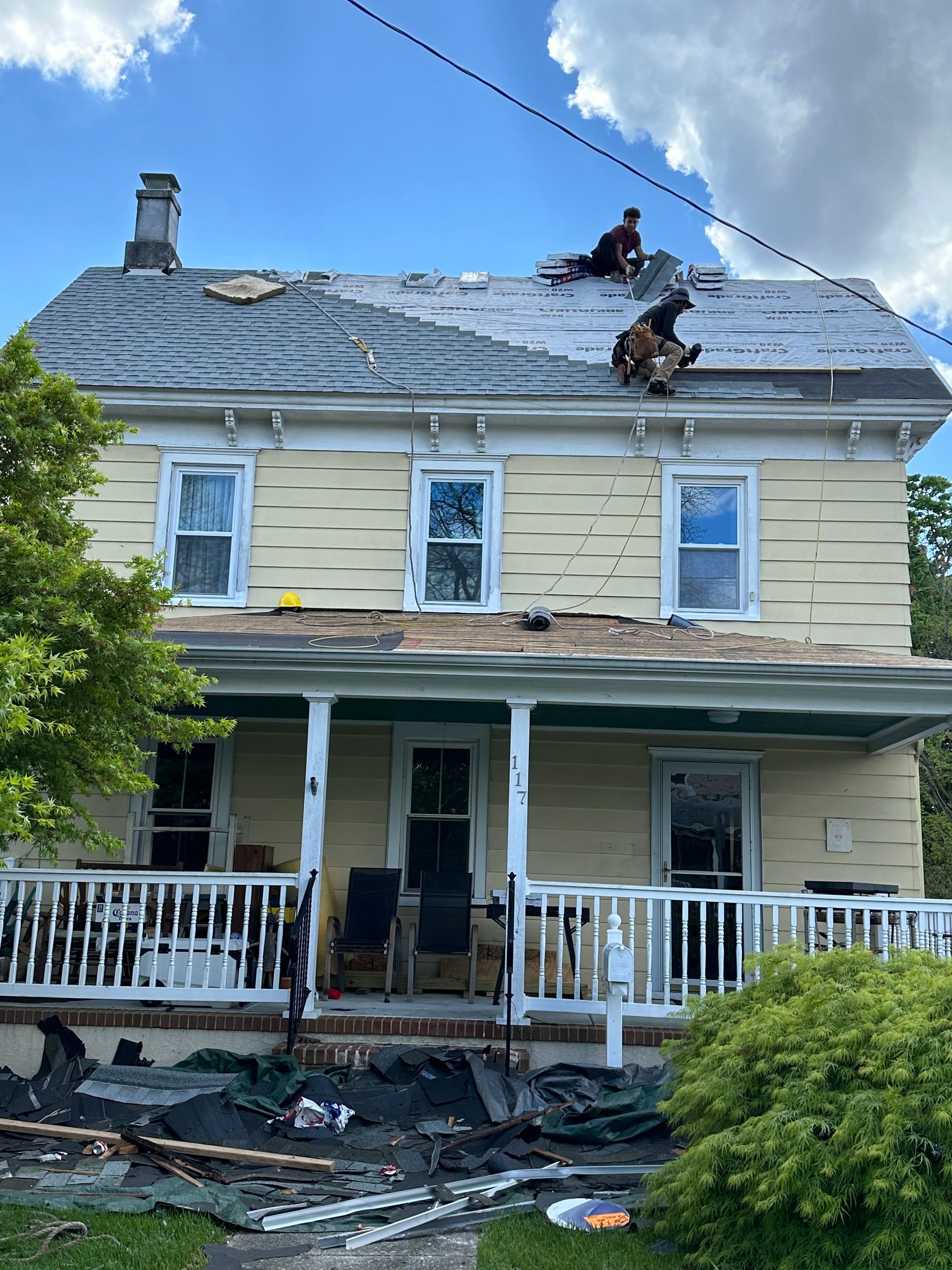 Damaged two-story house with roof and porch damage after a storm, debris scattered in front yard