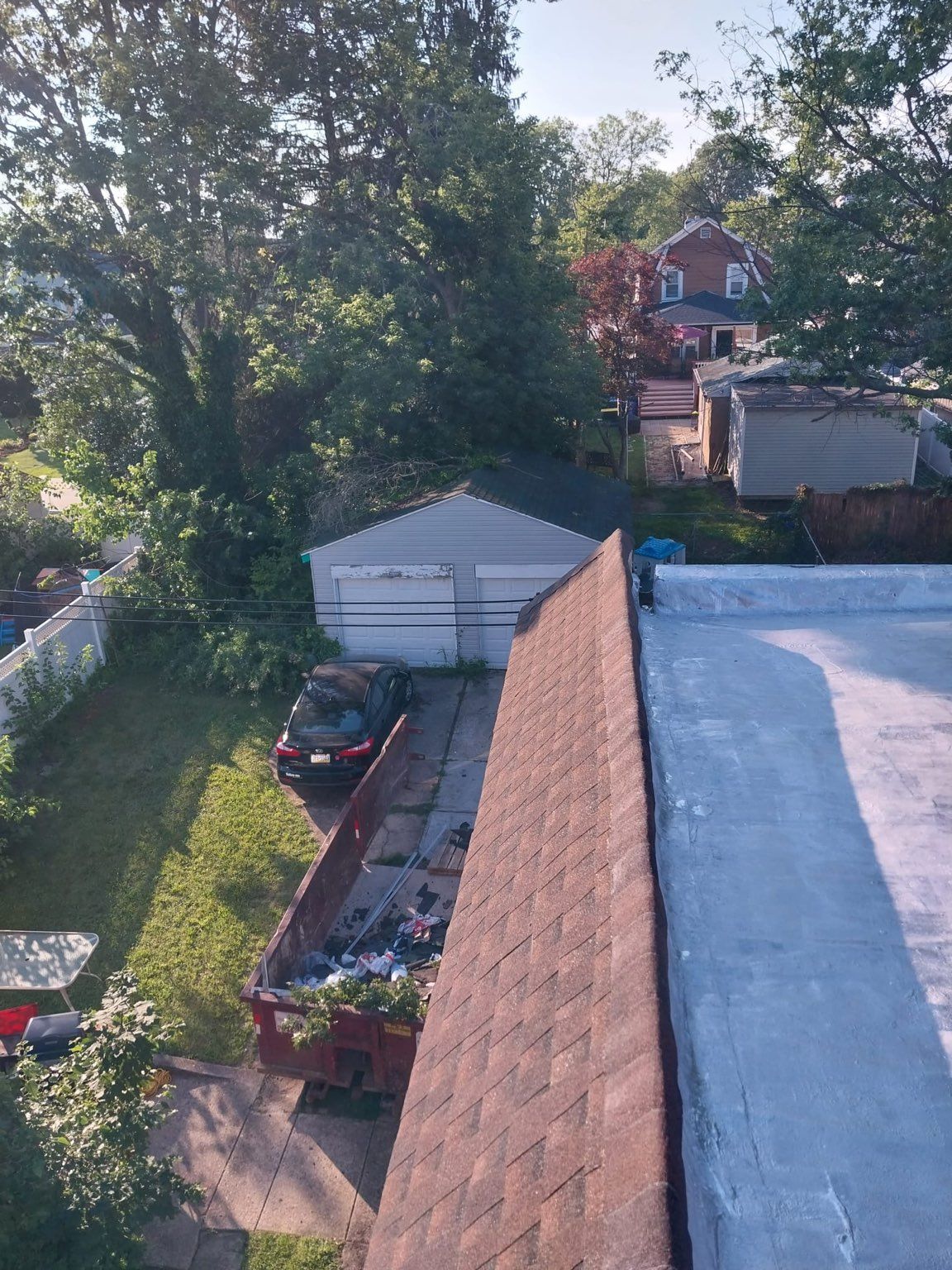 Rooftop view of a driveway, garage, and tree-lined yard with a parked car and neighboring houses in the background