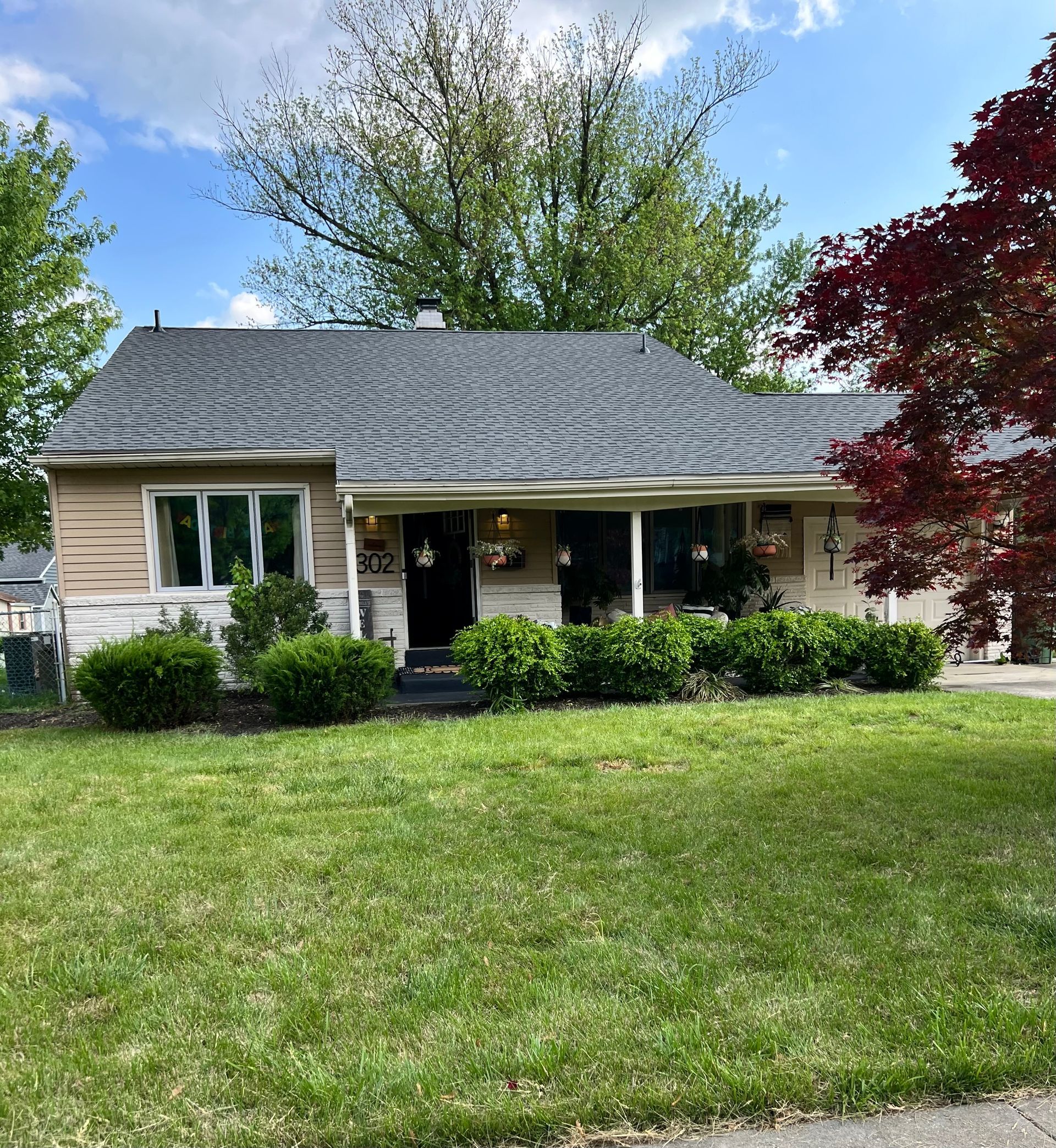 Single-story house with gray roof, front porch, green lawn, and trees under a blue sky